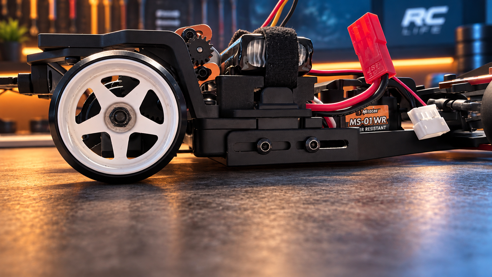 Close side view of the HPI chassis with a white wheel and the main chassis plate sitting higher over the workbench